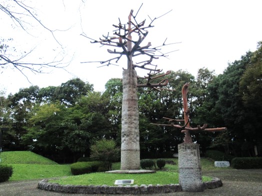 木々に囲まれた公園内に、幹は石造り、枝は鉄で出来た二本の木を模した抽象的な彫刻で、大きな彫刻が小さな石で囲まれた円形のこんもりした芝生の中央に立ち、小さな彫刻は枠の上に立ち並んでいる屋外アート作品の写真