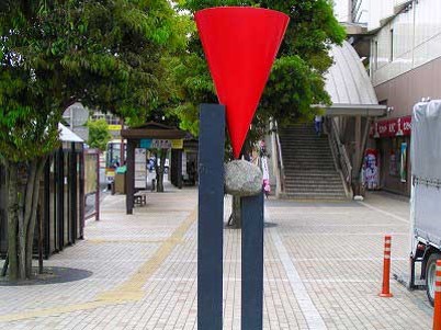 黒い二本の柱の上に、石と赤い逆さまの円錐が積み重なるように配置された、駅前広場に立つ現代彫刻の写真