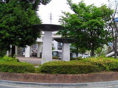 木々や生垣に囲まれた広場に設置された、異なる高さの二本の石柱の上に、黒い皿状の円盤が乗った、キノコのような形をした抽象的な屋外彫刻の写真