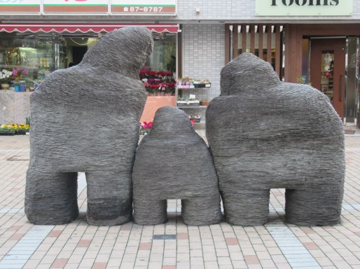 凹凸のある灰色がかった表面を持つ、親子を模したような三つの抽象的な石の彫刻が、商店街の歩道に寄り添うように設置されている写真