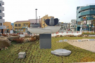 屋外の芝生広場の中心に、翼を広げた鳥と船を模した二つの石が、波打つような台座の上に配置されている彫刻の写真