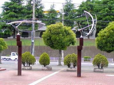 レンガ調の敷石が敷かれた広場に、赤茶色の柱の上で白い円弧状のオブジェが回転する一対の彫刻が設置されている写真