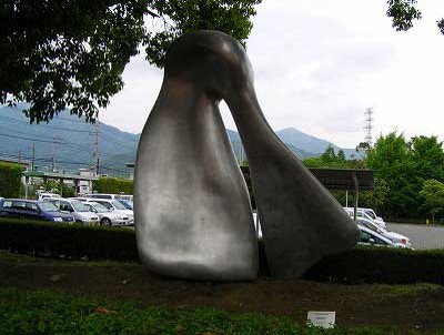 木々や生垣に囲まれた屋外に設置された、銀色に光る抽象的な形状で滑らかな曲線と光沢のある表面を持つ、2つの巨大なオブジェが上で円形になりつながっている金属製彫刻の写真