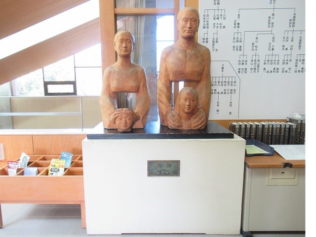 冊子や本が並んだ室内に、子供の頭や両肩に手を置いた親子を表すような木製の抽象的な像が二体、白く四角い台座の上に置かれている写真