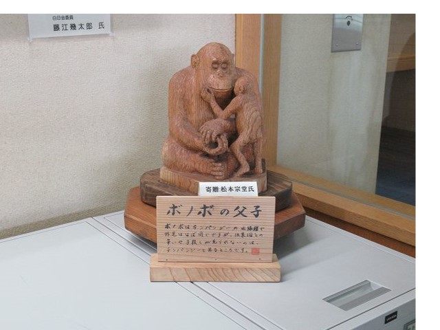 「ボノボの親子」と書かれた木製の看板が設置された屋内に設置された、大きなボノボが子供のボノボを優しく抱きかかえる様子を表現している、親子を描いた木製の彫刻の写真