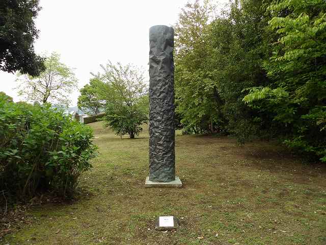 屋外の木々に囲まれた芝生の上に立つ、黒っぽい円柱形の彫刻で、表面はごつごつとした岩肌のような質感を持つアート作品の写真