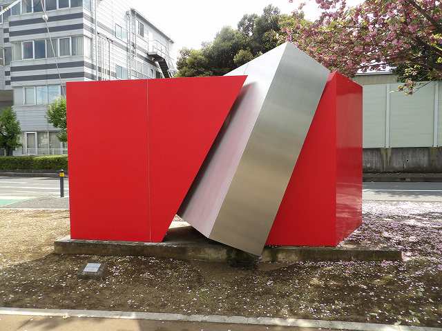 斜めに切り裂かれた赤い立方体の間に、銀色の直方体が突き刺さったような、ダイナミックな形状の現代彫刻が屋外に展示されている写真