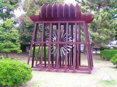 木々に囲まれた広場の中央に、赤茶色に塗装された木製の柱で組まれた立方体の枠の中に、鋭い歯車のような金属製のオブジェが設置されている写真