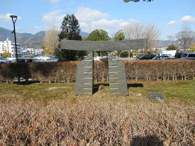 生垣に囲まれた広場に設置された、等間隔で横線が刻まれた二つの石の柱の上に、湾曲した一枚の板を載せたような鳥居を抽象化した石造りの屋外彫刻の写真