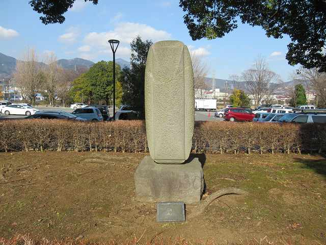 沢山の車が停まっている駐車場の傍の生垣に囲まれた広場に設置された、石の台座の上に石塔のような形をしたモニュメントが乗っている、抽象的な石造りの屋外彫刻の写真