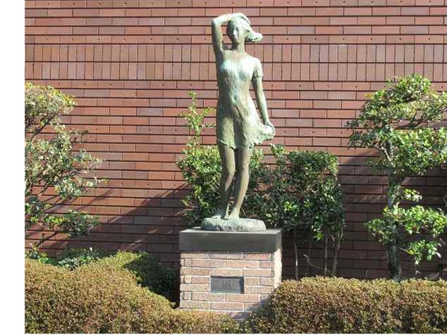 低木や生垣のあるレンガ造りの建物の前に、髪や服が風に吹かれなびいている薄い布をまとった女性が、右手を頭に置いて遠くを見つめている様子の全身像が、レンガでできた台座の上に立つブロンズ像の写真