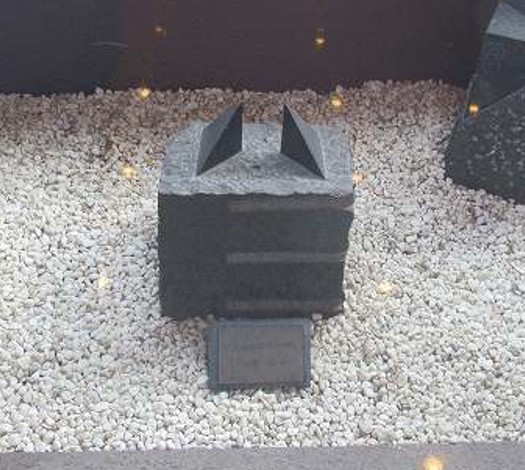白い小石を敷き詰めた中に、3箇所の横線が入った黒い立方体の石の上に、2つの三角錐が置かれた抽象的な室内彫刻の写真