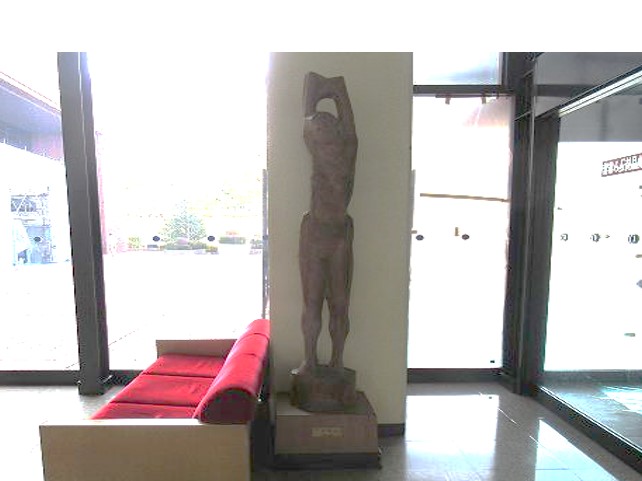 ロビーの赤いソファの横に腕を上げて何かを掲げるようなポーズをした、木製の男性の全身像が四角の台座の上に置かれている写真