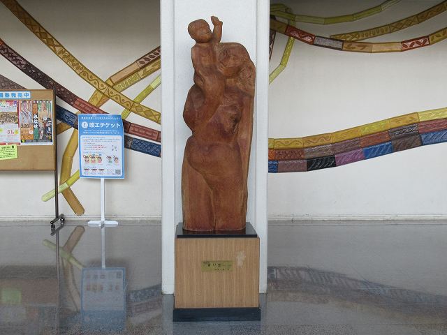 白い柱の前に設置された、右手を上げて空を仰いでいる小さな子供を右肩に乗せた母親の親子を象った木彫りの彫刻の写真