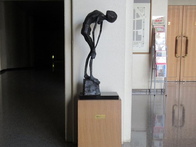 白い柱の前に設置された、前かがみになってうなだれる痩せた人物を象ったブロンズ像が、木製の台座の上に置かれている写真