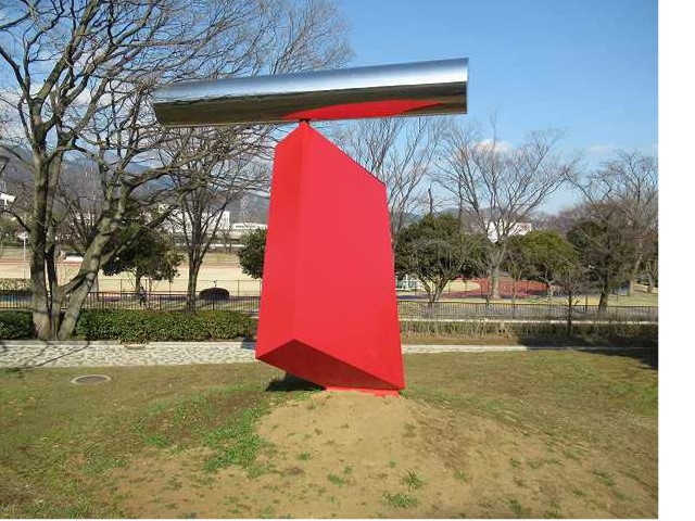 芝生広場の小高い丘に設置された抽象的なオブジェで、今にも倒れそうな斜めに設置された赤い多角形の土台の上に、銀色に輝く円筒形の物体が乗っている屋外彫刻の写真