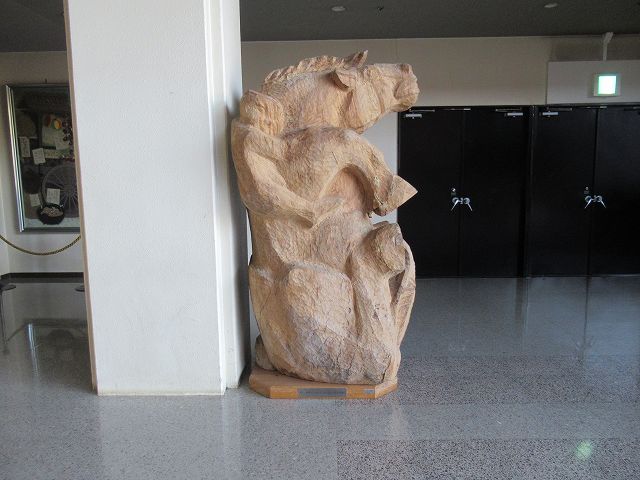 馬の背中に少年が抱きついている姿を力強く表現した、風合いのある木彫りの像が室内の白い柱際に置かれている写真