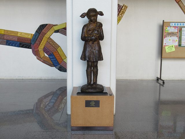 足元の台座に「愛」と記された、おかっぱ頭に二つ結びをした少女が、愛おしそうに人形を抱きかかえる姿を象った、ブロンズ製の彫刻が白い柱の前に設置されいる写真