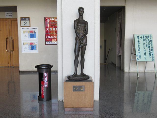 足元の台座に「少女」と記された、細身の女性が何かを手に持っているような、ブロンズ製の立像が屋内の白い柱の前に設置されている写真