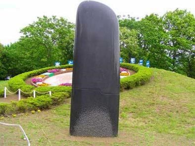 木々に囲まれた小高い広場の花壇や生垣で縁取られた芝生の上に立つ、黒く滑らかな表面を持つ、巨大な短刀のような形をした抽象的な彫刻の写真