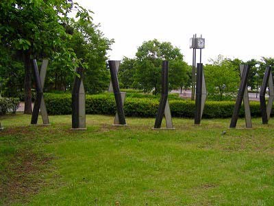 生垣や木々に囲まれた公園の芝生の上に、等間隔に配置された、黒と銀色の異なる素材が組み合わさり捻れたような形状をした複数のポールが芝生の上に並んでいる野外彫刻の写真