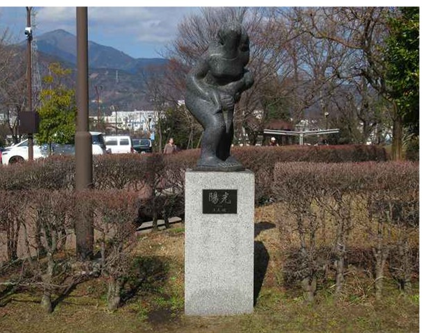 遠くに山並みが見える公園内の生垣の傍に立つ「陽光」と記された台座の上に、子どもを抱く親の姿を象った、滑らかな質感の抽象的な石造りの彫刻の写真