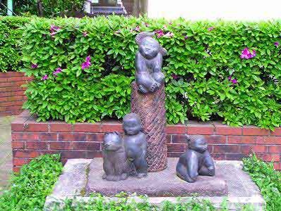 赤レンガ作りの花壇の中にピンク色の花が咲いた生垣を背景に、円柱の上に一体、その下に三体の幼い子どもや動物を思わせるブロンズ像が配置された屋外彫刻の写真