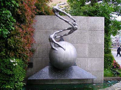 3人の人物が手を取り合い、丸い地球儀のような物体の上で弧を描くように連なっている、金属製の抽象的な屋外彫刻の写真