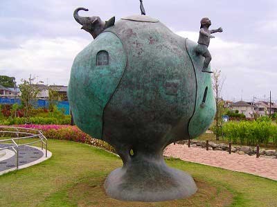 通路沿いの芝生広場に設置された、地面に土台とくびれのある大きな円形の地球のような形をし、表面に小さな窓がある抽象的な青銅色の彫刻の上に、ゾウや両手を前に伸ばしている躍動感のある子どもが配置された、物語性のある屋外彫刻の写真