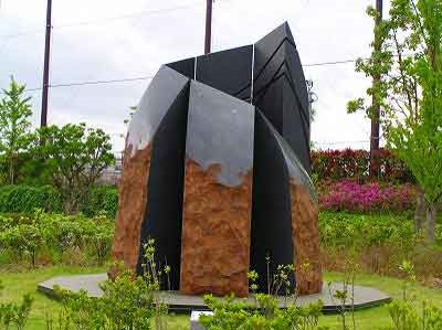 フェンスに囲まれた緑豊かな広場に設置された、長方形の表面がゴツゴツした様な茶色い石材が組み合わされた上から黒い液体を流したように、右側から左上に向かって螺旋状に配置された金属板が頂上に向かって尖るようにデザインされた、抽象的な屋外彫刻の写真