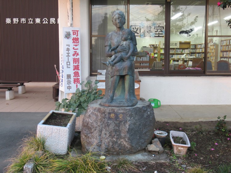 ガラス窓に図書室と書かれた建物の前に、「慈愛」と記された巨大な自然石の台座に、座っている母親が子どもを抱きかかえる姿を象ったブロンズ像が設置されている、公共施設の入り口で撮影された写真