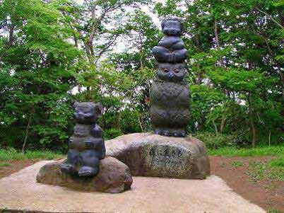 緑豊かな木々を背景に、前後に並んだ不揃いな石の土台の上に、頭の上に腕を組んだ猿が乗った大きなフクロウ、もう一つの石の上にクマのような動物が座っている様子を表現した屋外彫刻の写真