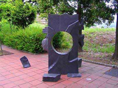 木々に囲まれた通路上に、真ん中に丸い穴が空いた黒い一枚の石板が、割れた破片とともにタイル敷きの地面に配置されている抽象的な屋外彫刻の写真