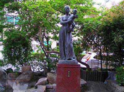 周囲を緑の木々で囲まれている屋外に、赤ちゃんを優しく抱く母親の姿を象ったブロンズ像が、赤い台座の上に立っている彫刻の写真