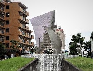 アパートやビルに囲まれた円形の広場に、背景のマンションが透けて見える薄い金属メッシュで作られた波打ちながらねじれたような抽象的なモニュメントが立っている写真