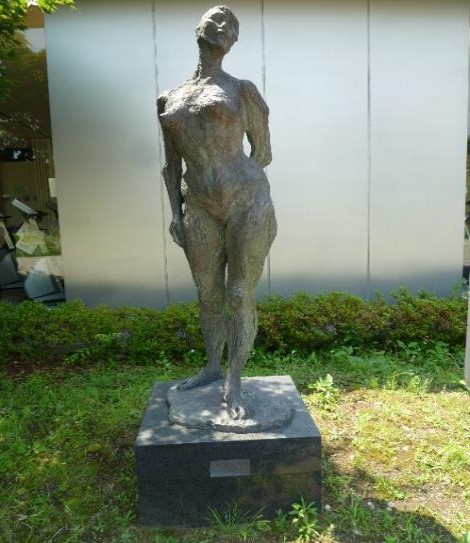 頭部や腕が抽象化された、痩せた女性の裸体を表現したブロンズ製の立像が、屋外の台座の上に設置されている彫刻の写真