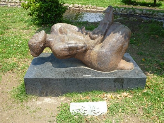 水が流れる水路の傍の広場に、母親が仰向けになりながら子どもを抱きかかえる姿を象った、ブロンズ製の抽象的な彫刻が、屋外の黒い台座の上に設置されている写真