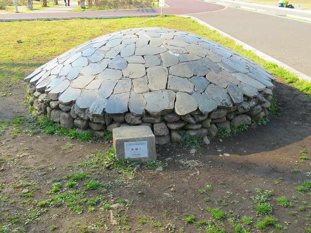 ところどころ草が生えた通路の角に「集積1」と書かれた石のプレートが置かれ、不揃いな石がドーム状に積み重ねられた屋外のオブジェの作品の写真