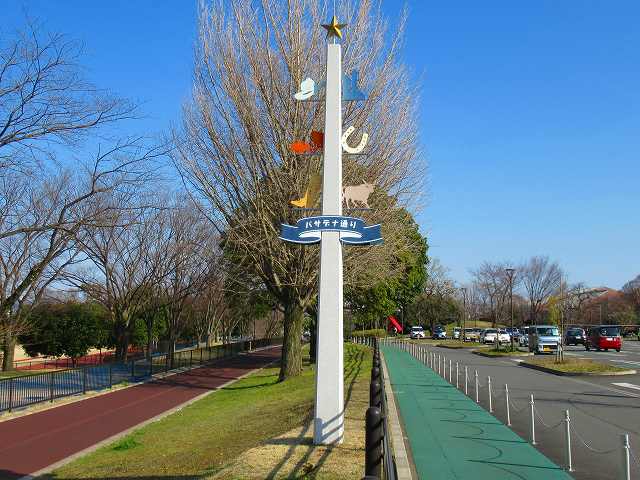 通路と駐車場に面した木々が立ち並ぶ公園に、上に向かって細くなっている中央部分の青いリボンに「パサデナ通り」と書かれ、頂上に星、馬蹄やウエスタンブーツなど様々な飾りがついた白い柱が立っている写真