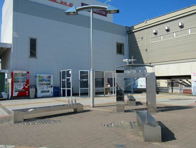 対曲線状に設置された4つの銀色に輝く金属製のベンチの中央に、門の様な形のコの字型のオブジェがあり、傍に空中に浮かんでいるようなヤジロベエの様な形をしたオブジェが特徴的な抽象的な彫刻が、駅前の広場に設置されている写真