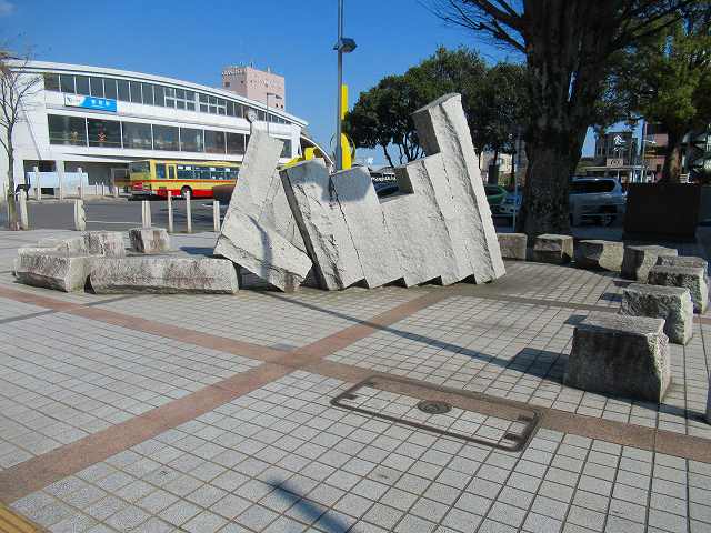 背景に駅舎が見える駅前広場の舗装された地面に並べられた、倒れそうに重なって斜めに立っている高さの違う5枚の砕けたコンクリートブロックのような抽象的な石が、L字型の石に支えられて立っている彫刻の写真