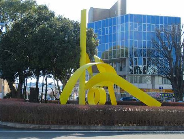 背景に全面ガラス張りのビルが立ち大きな木々や生垣に囲まれた円形の敷地の中央に、明るい黄色のリボンが絡み合ったような抽象的な現代彫刻が設置されている写真