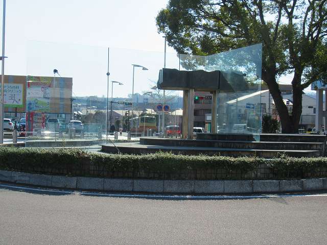 背景に一本の大きな木が立つロータリーの階段状の中央にガラス板がL字形で配置され、その間に鳥居の様な形をした重厚な黒御影石の物体が挟まれている抽象的な屋外彫刻の写真