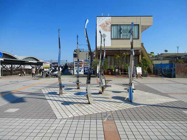 複数の光沢のある細長い金属製のポールが、不規則に地面から突き出ている抽象的な彫刻が広場に設置されている写真