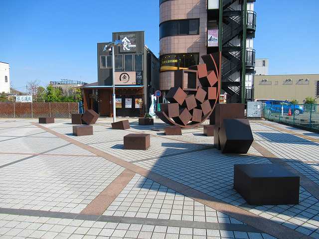 茶色い金属製の複数の直方体や立方体が、中央の巨大な半円形のオブジェから飛び出したように広場に配置された抽象的な屋外彫刻の写真