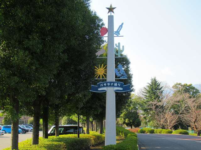 駐車場と並木道沿いの間に、上に向かって細くなっている中央部分のブルーの帯に「パサデナ通り」と書かれ、頂上に星、イチゴやサボテンなど様々な飾りがついた白い柱が立っている写真