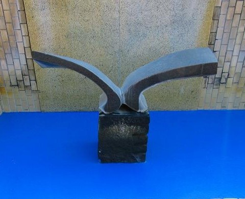 四角い黒い台座の上に、羽を広げた鳥を抽象的に表現したような、灰色でざらついた質感の彫刻が展示されている写真