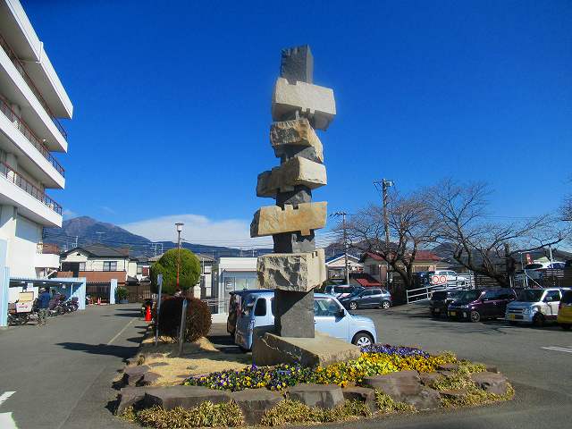 白い建物が建ち沢山の車が停まっている駐車場の敷地内に、窪みのあるいくつかの石が不安定に積み重ねられたような、高くそびえる抽象的な石の彫刻が、花壇の中心に設置されている写真