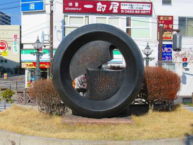 背景にコンビニが映る通りに面した小さな植え込みに、中心に穴が空いた黒いドーナツ型に、一部を切り取った金属板を組み合わせたような抽象的な現代彫刻の写真