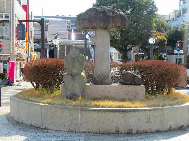 歩道の中央にある円形の植え込みにある長方形の土台に、顎に両手を置き物思いにふけっているような人物の石像と、雲のような形をした柱の彫刻、そして動物の彫刻が設置されている写真